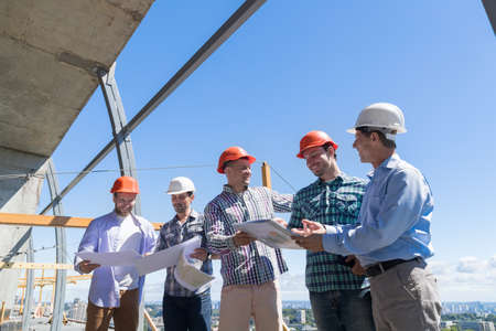 Group Of Builders In Hardhat Discussing Project Blueprint With Architect On Constuction Site Working Together Teamwork Conceptの写真素材