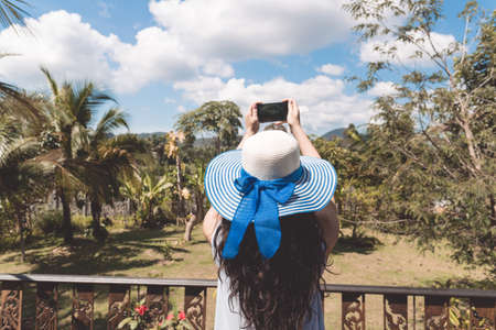 Young Woman In Hat Make Photograph Of Tropical Forest Landscape From Balcony Or Terrace Back Rear Viewの写真素材