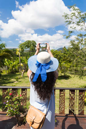 Rear View Of Young Woman In Hat Taking Photo Of Tropical Forest On Smart Phone From Balcony Or Terraceの写真素材