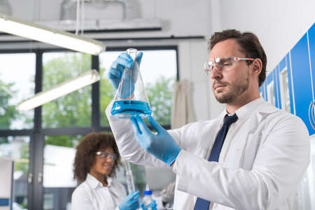 Serious Scientist Looking At Flask With Blue Liquid In Laboratory Over Group Of Scientific...