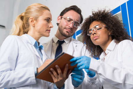 Group Of Scientific Workers Taking Notes Making Research In Laboratory, Mix Race Team Of Scientists Writing Results Of Experiment Or Diagnosis In Tablet In Labの写真素材