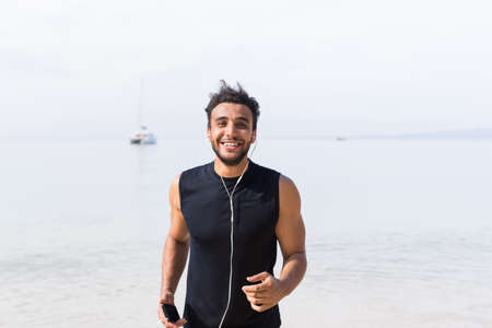 Portrait Of Young Hispanic Man Runner On Beach Holding Cell Smart Phone And Listening Music While Jogging On Seaside Male Happy Smiling Latin Sport Healthy Lifestyle Conceptの写真素材