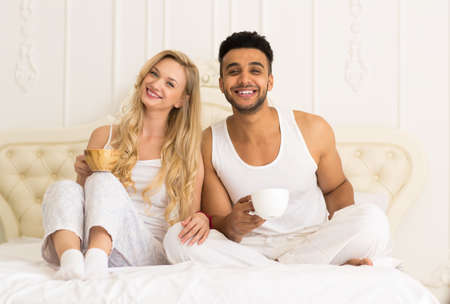 Young Couple Drink Coffee Orange Juice Sitting In Bed, Happy Smile Young Hispanic Man And Woman Lovers Hold Cups Glasses Bedroomの写真素材