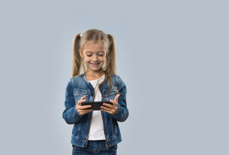 Beautiful Little Girl Using Cell Smart Phone Happy Smiling Wear Jeans Coat Isolated Over Gray Backgroundの写真素材
