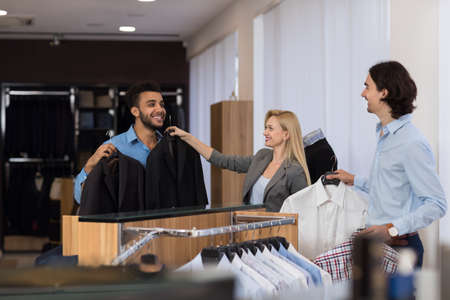 Female Shop Assistant Help Young Handsome Business Man Choose Suit In Clothes Shop, Two Businessmen...