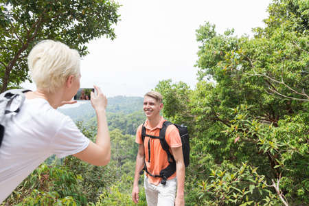 Girl Taking Photo Of Guy With Backpack Posing Over Mountain Landscape On Cell Smart Phone, Trekking Young Man And Woman Group On Hike Tourists Adventure Activityの写真素材