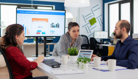 Three colleagues engaged in a productive discussion around a table in a bright, contemporary office space, with a large screen displaying charts.の素材