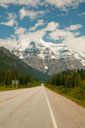 Stretch of road leading to snow covered Mount Robson against blue sky.の写真素材