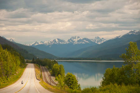 View of highway and majestic snow covered mountains in the distance with a calm lake in the foreground.の写真素材