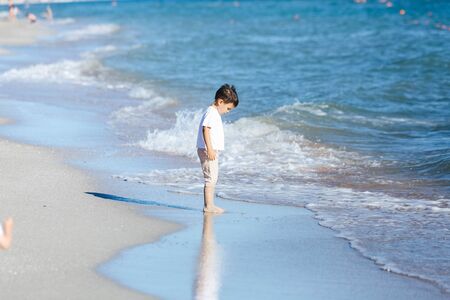 a child in a white t-shirt and light shorts stands on the beach and looks at the wave.の写真素材