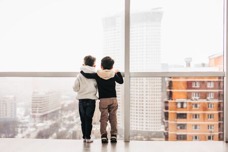 Two little boys are embraced standing and looking through a panoramic window to the street.  Stay at home.の写真素材