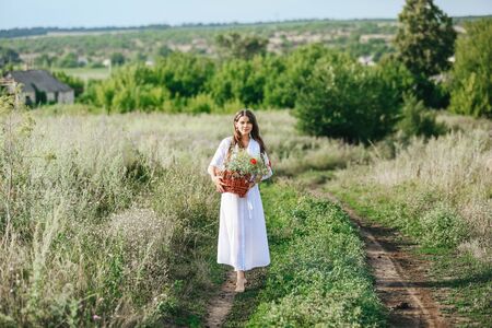 The young beautiful girl in a white dress goes in the field and holds a basket with wildflowers. Summer story.の写真素材