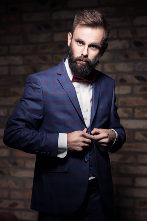 man with beard in suit staying by the brick wall and buttoning; charismatic bearded guy man with beard in suit staying by the brick wall and buttoning; charismatic bearded guy