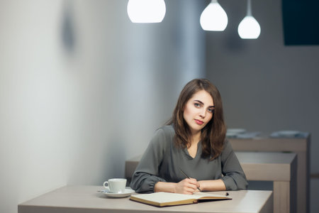 young adult female has coffee break in cafe and writing notes in diary or notepad;