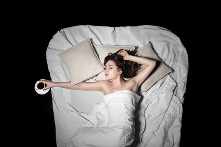 young female lying in bed and keeps coffee, view from above;の写真素材