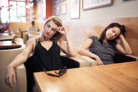 two tired females sitting in cafe and resting; weary girls have coffee break after hard working day;