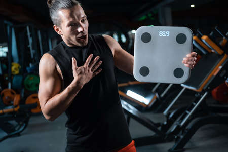 shocked and frustrated man weighing with scale in gym after dietingの写真素材