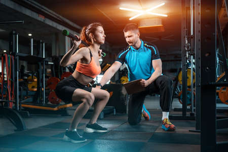 young female workout and exercising barbell squat in gym with personal trainerの写真素材