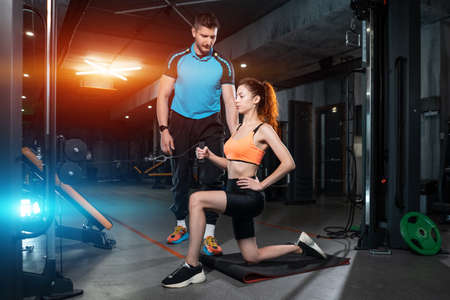 young woman exercising single arm row kneeling with personal trainer man in gymの写真素材