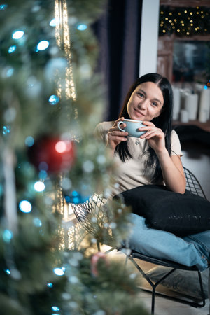 young female drink coffee and rest with cup by new year christmas tree at homeの写真素材