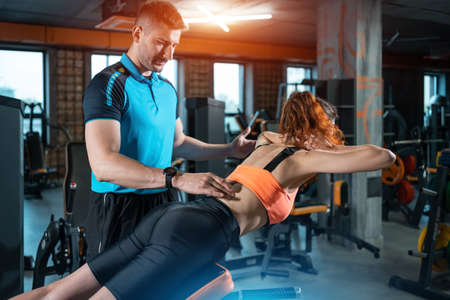 young woman exercising lower back hyperextension with personal trainer in gymの写真素材