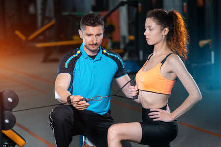 young woman exercising single arm row kneeling with personal trainer man in gymの写真素材