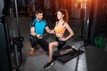 young woman exercising single arm row kneeling with personal trainer man in gymの写真素材