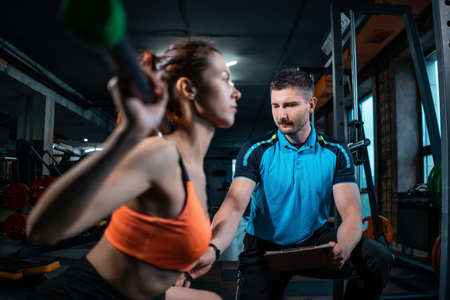 young female workout and exercising barbell squat in gym with personal trainerの写真素材