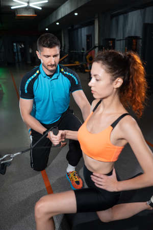 young woman exercising single arm row kneeling with personal trainer man in gymの写真素材
