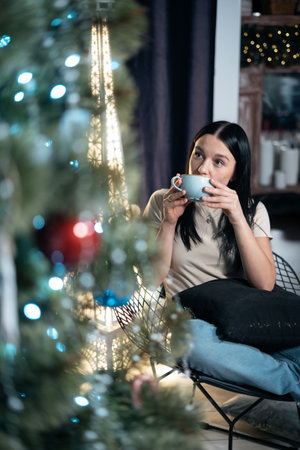 young female drink coffee and rest with cup by new year christmas tree at homeの写真素材