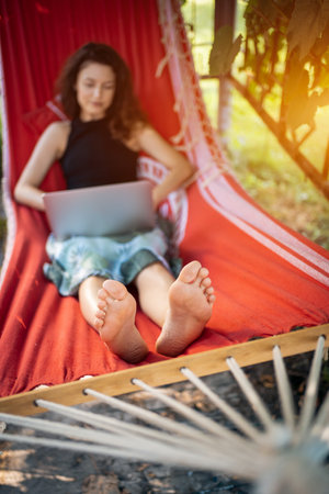 focused young female typing on laptop while lying in red hammock outsideの写真素材