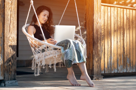 young female rest on macrame swing chair with laptop at country house terraceの写真素材