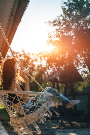 happy young female riding on macrame swing chair near country house outdoorsの写真素材