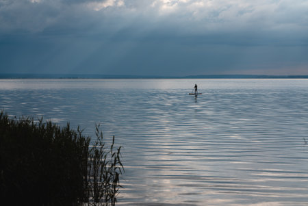 young man standing on sup and paddling on water of calm lake, tranquil sceneryの写真素材