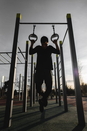 young athletic man exercising rings at fitness calisthenics workout and making pull-ups at street...