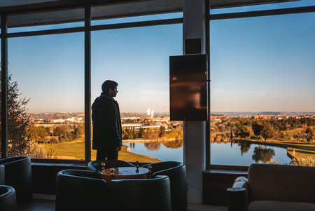 successful man by window of golf club looking at serene lake from modern indoor space during sunset