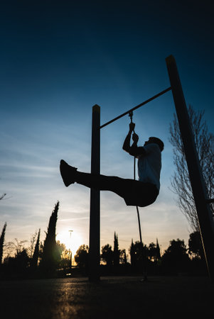 young athletic man exercising fitness calisthenics workout and making ring leg raises for abdomen abs muscles at street gym during sunset, body improvement and strength trainingの写真素材