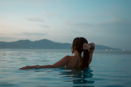 beautiful woman swimming in luxury pool at sunset flowing into sea, elegant vacation and tropical...