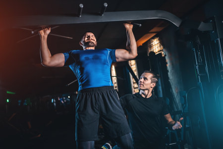 Male bodybuilder performing lat pulldown with personal trainer in gym, Man training back muscles under coach guidance during workoutの写真素材