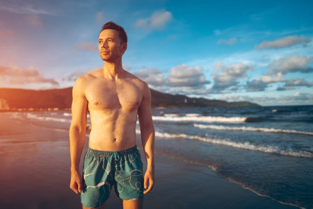 white man with athletic muscular body standing at beach at sunny sunset, male athlete walking at sea or ocean shoreの写真素材