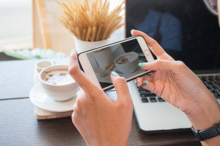 Woman taking a picture of a coffee cup with her smart phone while sitting at a coffee shop.