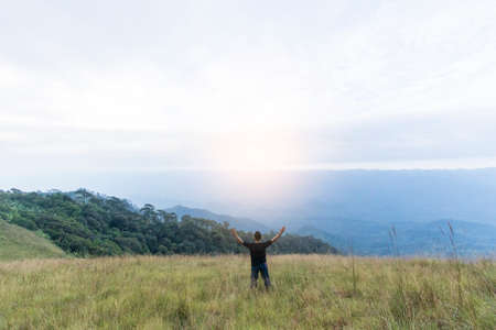 Happy young man standing with raised arms in green field on mountainsの写真素材