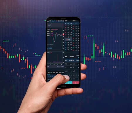 Trading concept. Business hand holding phone and touching screen on smart phone over blur stock chart market backgroundの写真素材