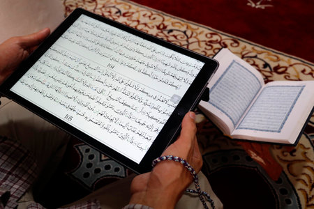 Muslim reading a digital Quran on a tablet.In the background a traditional paper Arabic Holy Quran.のeditorial素材