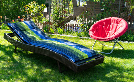 A blue and green colored sun lounger and a red round red folding moon chair in a green garden standing in the sun during spring time..の写真素材