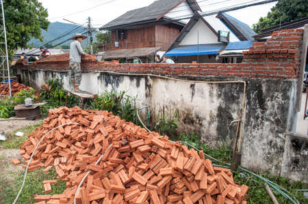 Pai, Thailand - November 26, 2014: A young man with a hat on a clear sunny day is building a wall from a red brick building. Thai street of a small town in the north of the country.のeditorial素材