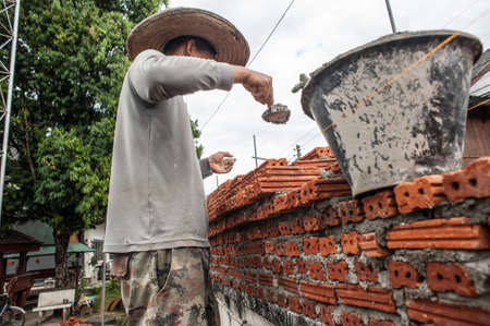 Pai, Thailand - November 26, 2014: A young man with a hat on a clear sunny day is building a wall from a red brick building. Thai street of a small town in the north of the country.のeditorial素材