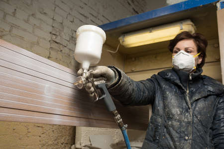 Saint-Petersburg, Russia - March 17, 2016: Carpenter painter paints the furniture board with a spray gun on the production line small wholesale furniture factory on the background of the air intake. Protective face mask.のeditorial素材