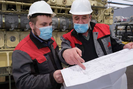 Saint Petersburg, Russia - April 24, 2020: Two engineers are discussing design ideas with blueprints in their hands against the background of a super powerful diesel engine in the workshop the plantのeditorial素材