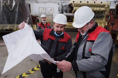 Saint Petersburg, Russia - April 24, 2020: Two engineers are discussing design ideas with blueprints in their hands against the background of a super powerful diesel engine in the workshop the plantのeditorial素材